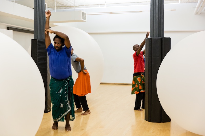 Three black men in primary colors and dance meditatively around large white orbs